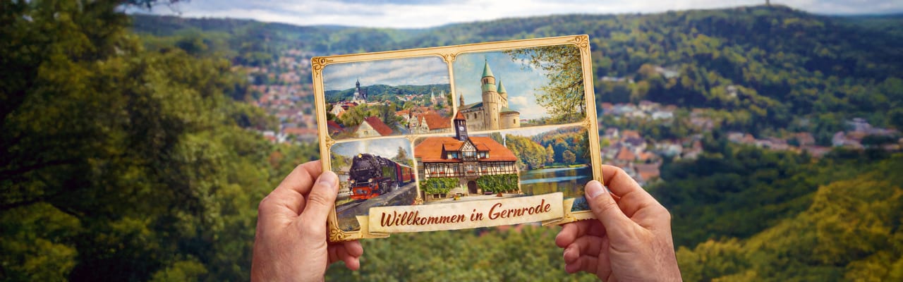 Hände halten eine Postkarte mit Motiven aus Gernrode vor einer Harzlandschaft